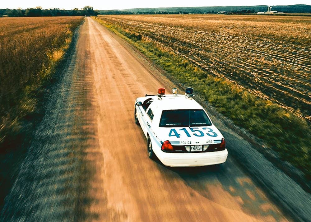 Drone footage capturing a police car speeding down a rural road amidst fields, en route to a crime scene for a TV show.