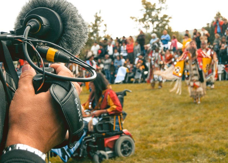 Close-up of a video camera filming First Nations individuals dancing at a Pow-Wow gathering.