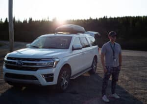 Richard Duquette, the fixer from Films.Solutions, posing beside a large white SUV with a sunset behind a fir tree forest at the entrance of Labrador City
