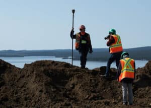 Mining specialist standing on a dirt pile with a cinematographer filming and director overseeing, against a backdrop of a lake in Labrador City