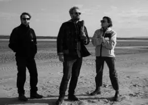 Black and white image of the cinematographer, liaison officer, and director posing fashionably on a sandy beach next to a river