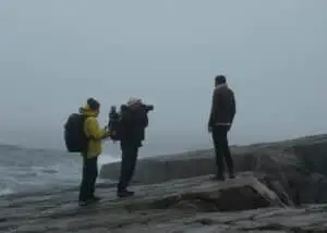 Director and cinematographer filming Terrence, an artist and seaweed specialist, on a vast flat rock with the harsh sea in the background, in Grate Cove, Newfoundland