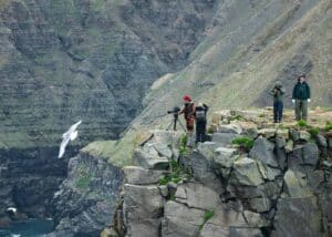 Film team near a cliff, preparing to film Luka, with Bird Rock, hosting thousands of nesting birds, in the background