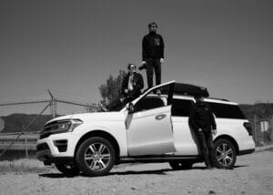 Black and white photo of the film team posing fashionably around and atop a white SUV during their filming adventure