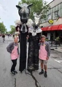 Two participants from 'Race Across the World 3' posing with vibrant mascots in the lively streets of Montreal.